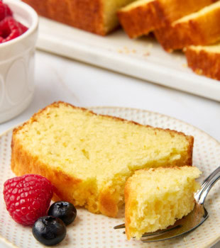 a slice of yogurt cake on a plate with a bite on a fork; more cake and fresh berries partially visible in the background