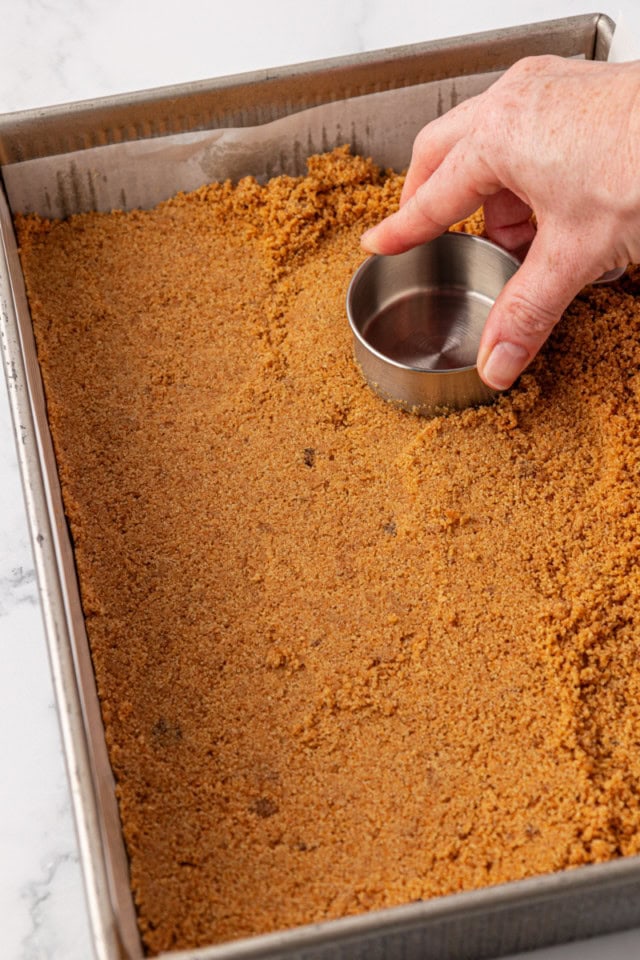 graham cracker crust being pressed into the bottom of a 9x13 baking pan
