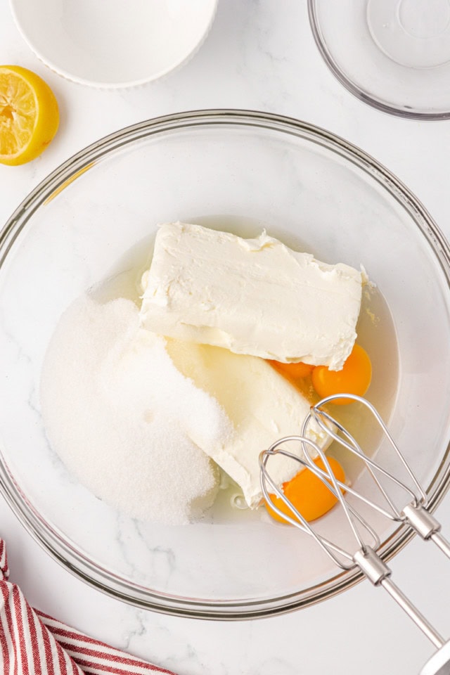 overhead view of cream cheese, sugar, eggs, and lemon juice in a glass mixing bowl