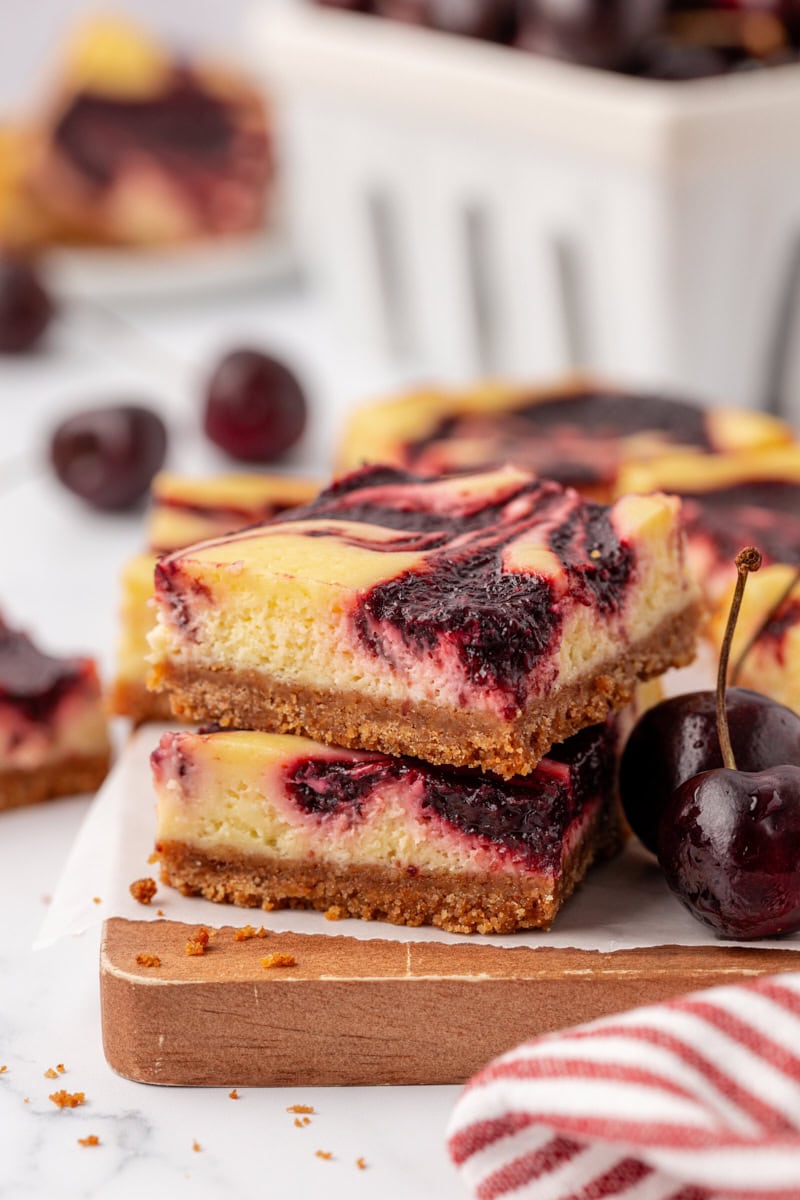 two cherry cheesecake bars stacked on a wooden board with fresh cherries in the background