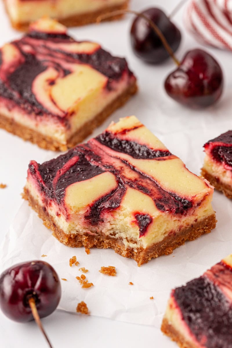 a cherry cheesecake bar with a bite missing, surrounded by more bars and fresh cherries