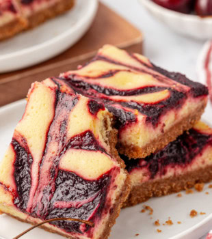 a cherry cheesecake bar leaned against a stack of two more bars; fresh cherries and more bars partially visible in the background