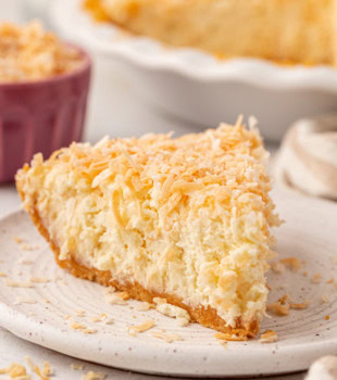 a slice of no-bake coconut cheesecake on a plate with remaining cheesecake and a bowl of toasted coconut in the background