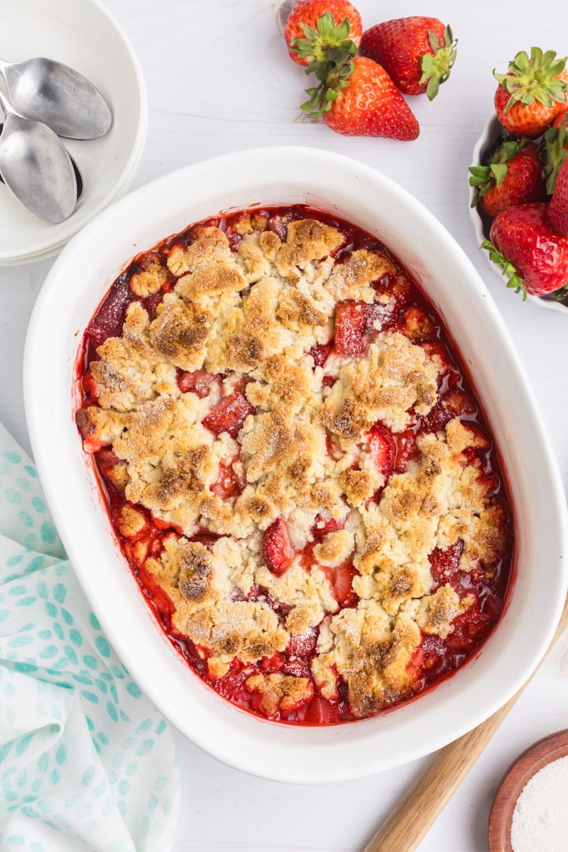 overhead view of freshly baked strawberry cobbler surrounded by fresh strawberries, bowls, and spoons for serving