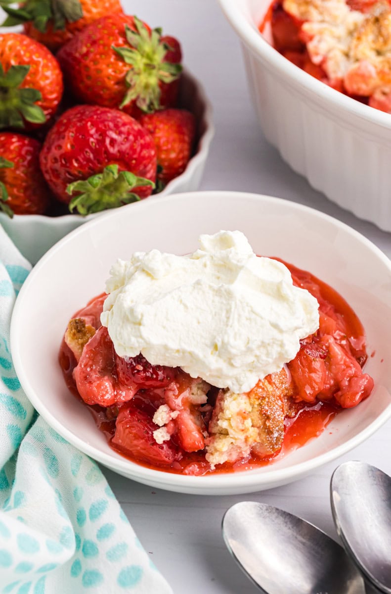 strawberry cobbler topped with whipped cream in a white bowl; remaining cobbler and fresh strawberries partially visible in the background