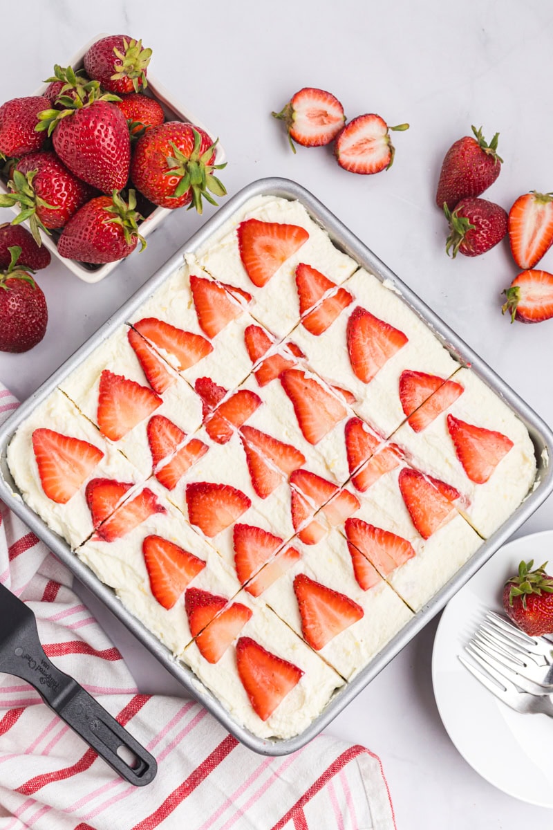 overhead view of strawberry tiramisu cut into 12 servings in the pan, surrounded by more strawberries, plates, and forks