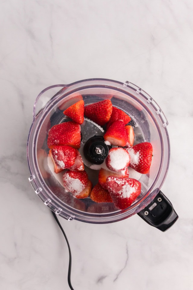 overhead view of strawberries, sugar, and lemon juice in the bowl of a food processor