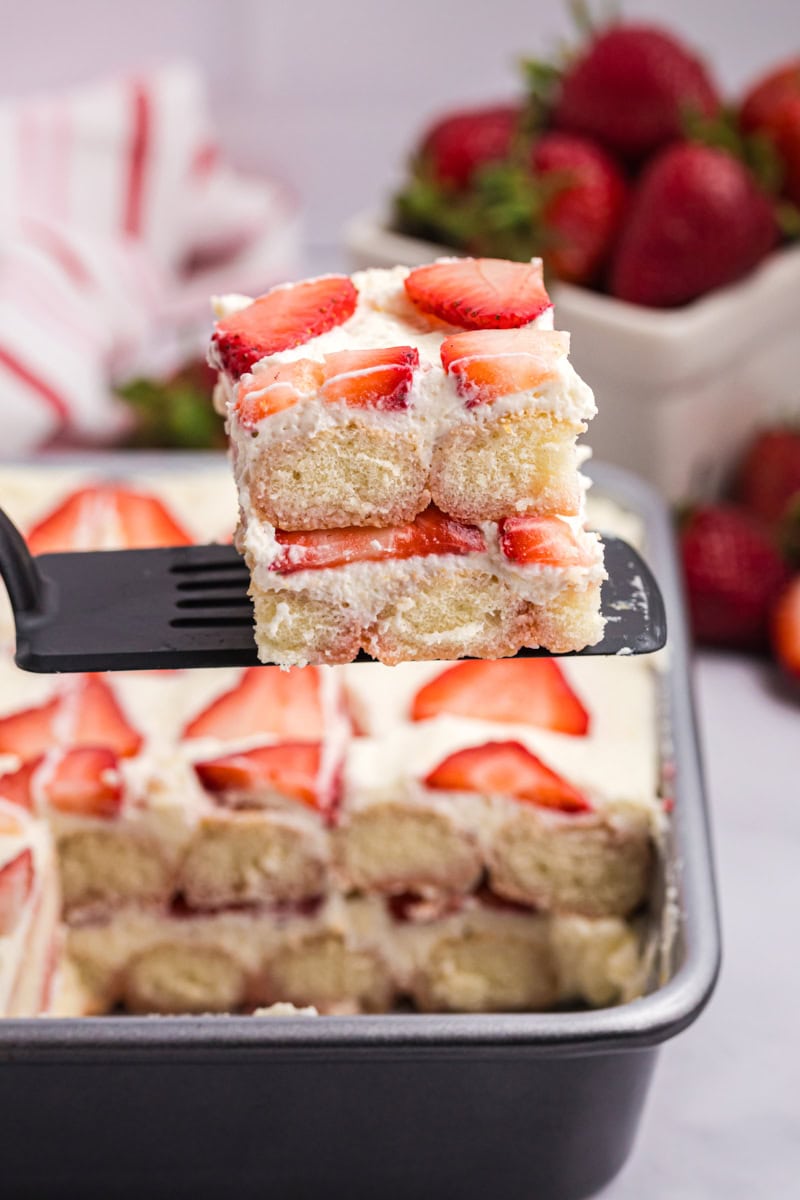 a slice of strawberry tiramisu on a spatula being lifted out of the pan with the remaining tiramisu