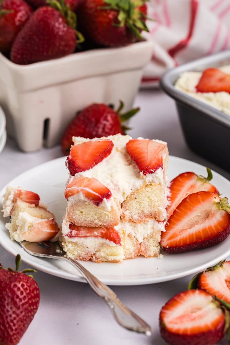 a slice of strawberry tiramisu on a white plate with the remaining tiramisu and a basket of fresh strawberries partially visible in the background