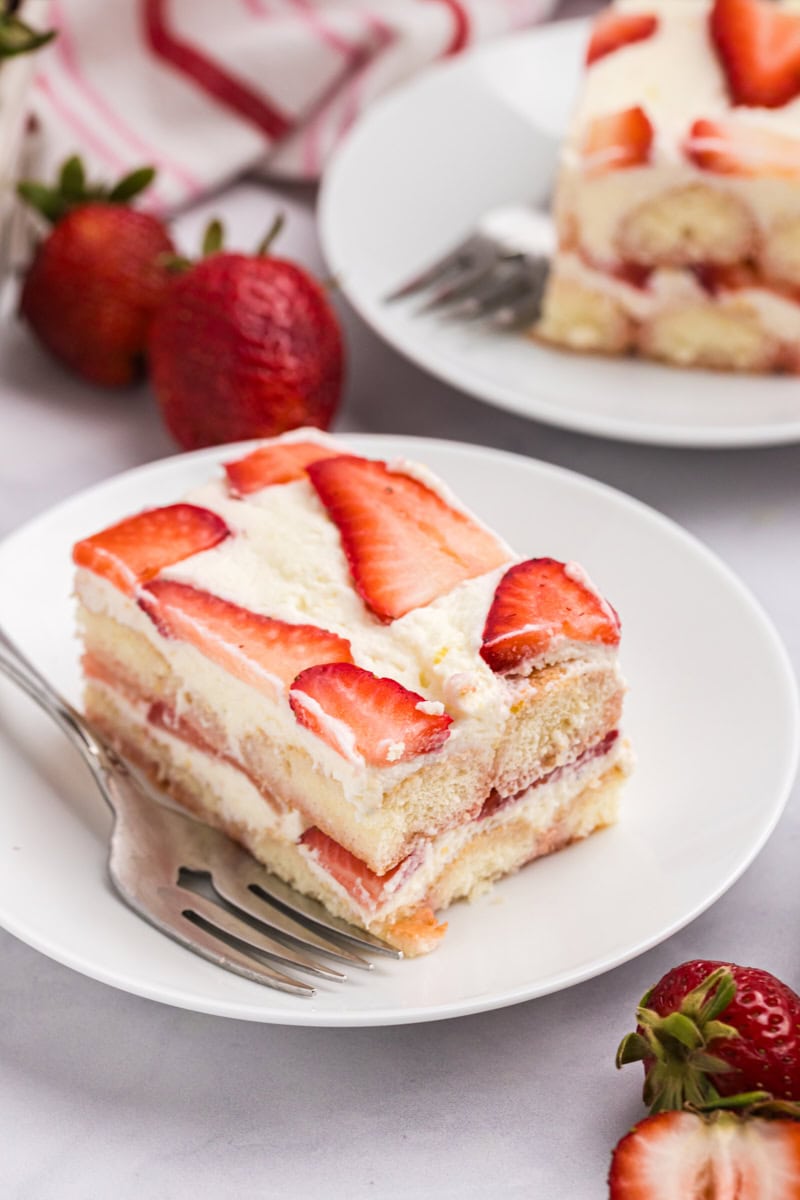 a slice of strawberry tiramisu on a white plate with fresh strawberries and another serving in the background