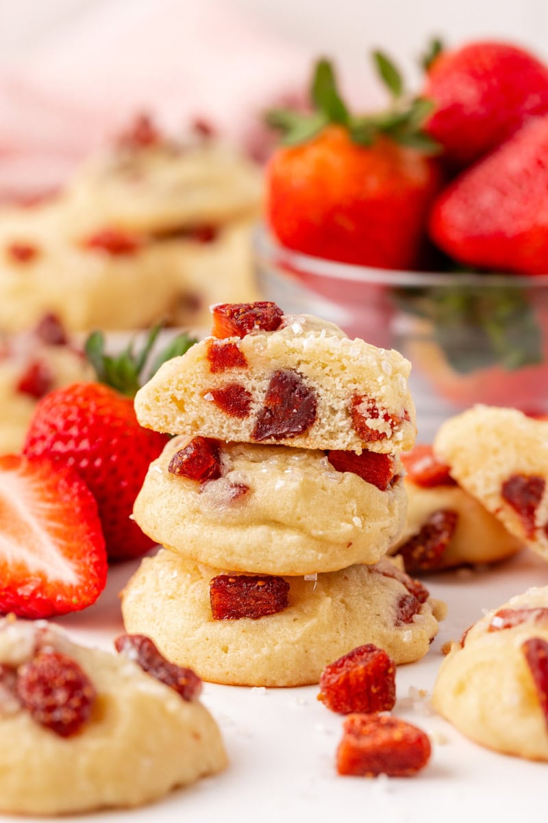 stack of three strawberry cream cheese cookies with a bite missing from the top cookie; more cookies and fresh strawberries in the background