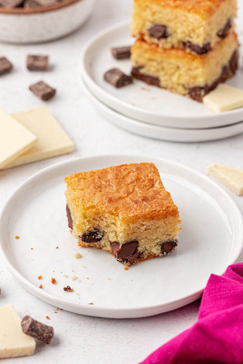 a white chocolate brownie with a bite missing, served on a white plate; more brownies in the background
