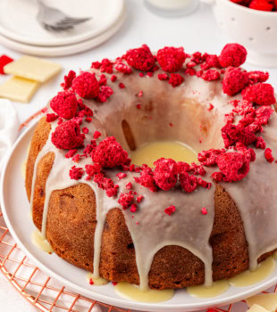 white chocolate raspberry Bundt cake on a white cake plate with plates and a bowl of fresh raspberries partially visible in the background