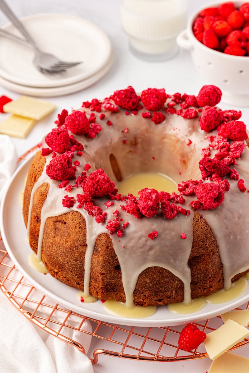 white chocolate raspberry Bundt cake on a white cake plate with plates and a bowl of fresh raspberries partially visible in the background