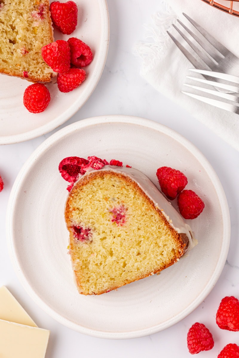 overhead view of a slice of white chocolate raspberry Bundt cake on a white plate with another slice, berries, forks, and white chocolate pieces partially visible