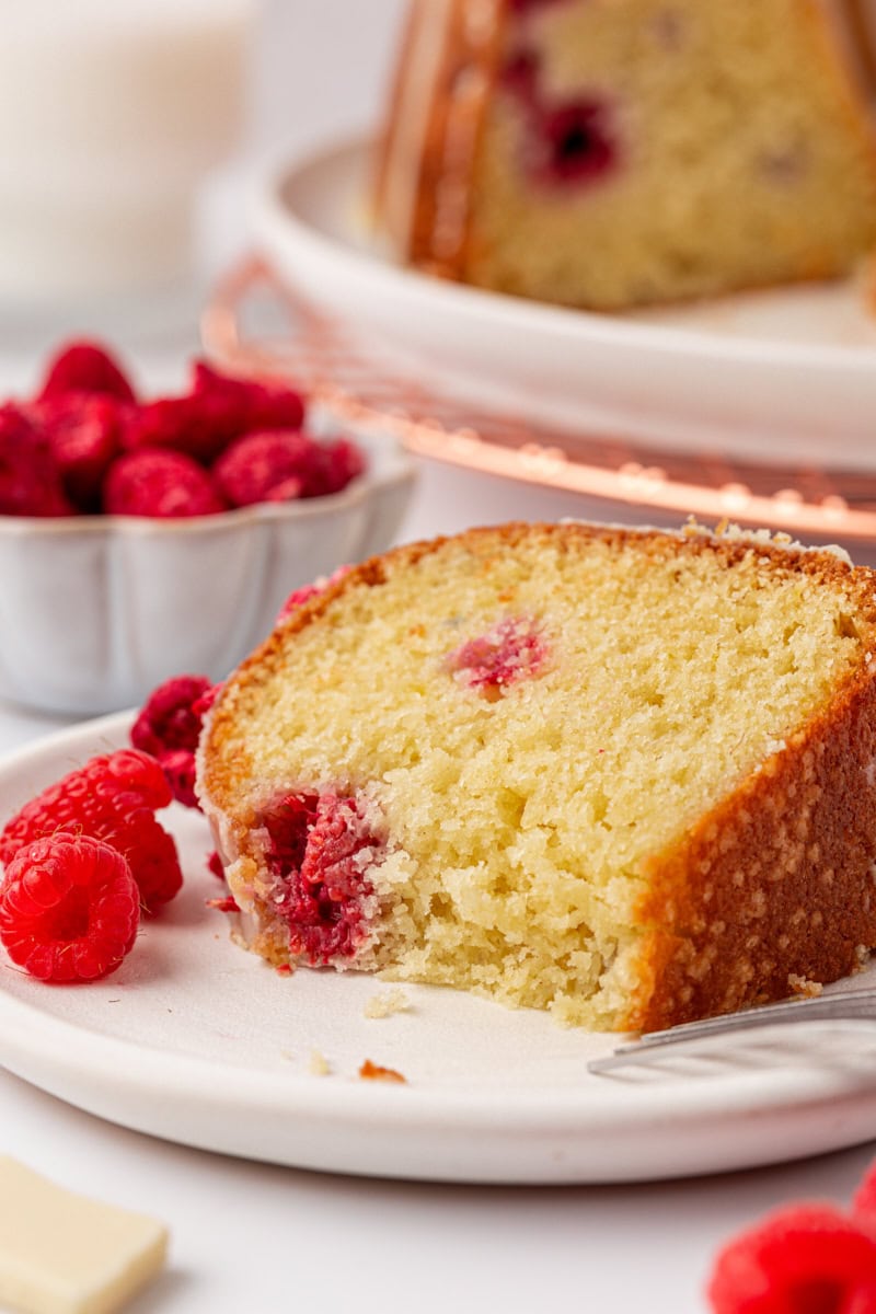 a slice of white chocolate raspberry Bundt cake with a big bite missing; more cake and a bowl of fresh raspberries in the background