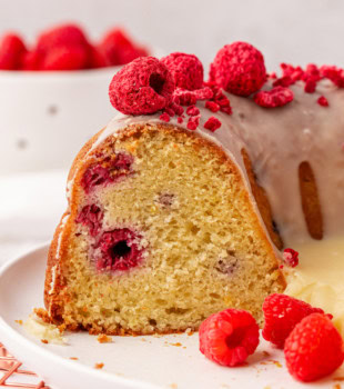 partially sliced white chocolate raspberry Bundt cake on a white cake plate with a bowl of fresh raspberries partially visible in the background