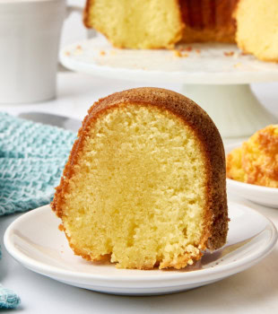 a slice of five flavor pound cake on a white plate with the remaining cake on a white cake stand in the background