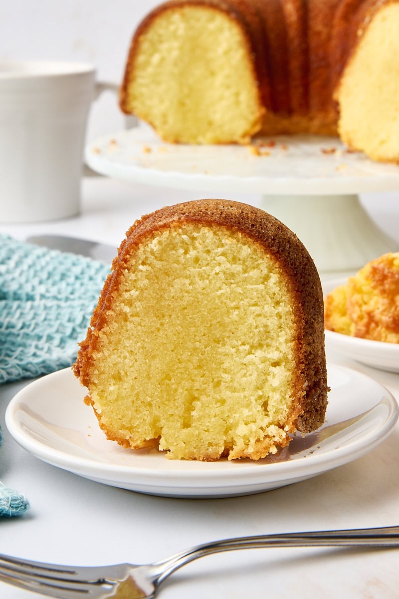 a slice of five flavor pound cake on a white plate with the remaining cake on a white cake stand in the background