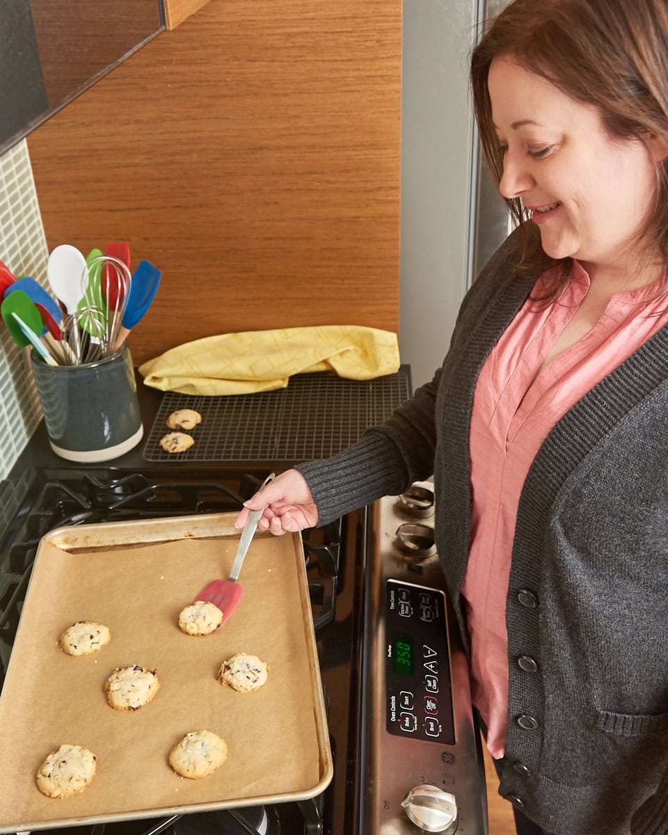 Jennifer of Bake or Break standing beside an oven removing freshly baked cookies from a parchment-lined baking sheet onto a wire rack