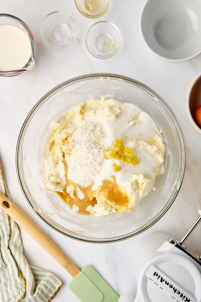 overhead view of sugar, vanilla, flour, and lemon zest added to beaten cream cheese in a glass mixing bowl