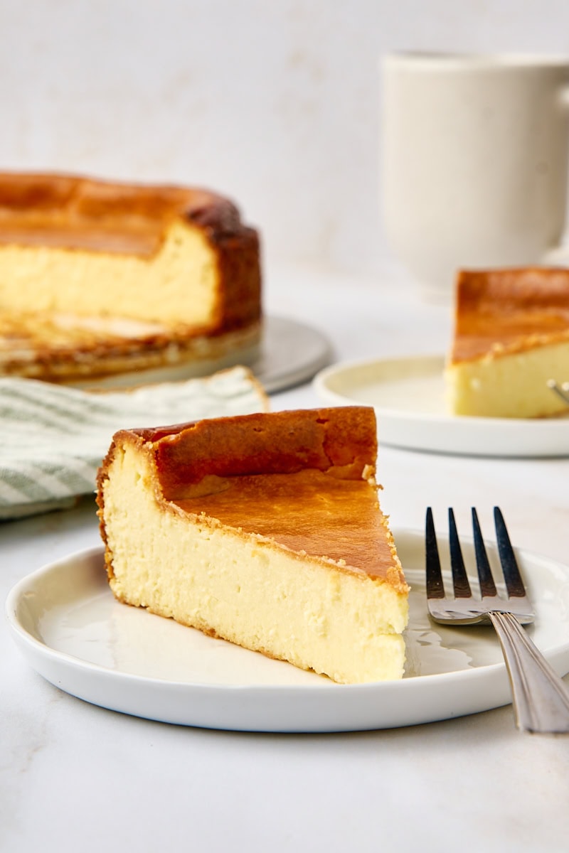 a slice of New York cheesecake and a fork on a white plate; more cheesecake and a coffee mug partially visible in the background