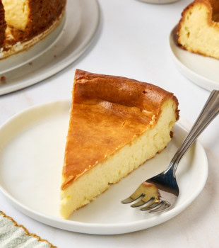 a slice of New York cheesecake and a fork on a white plate; more cheesecake partially visible in the background