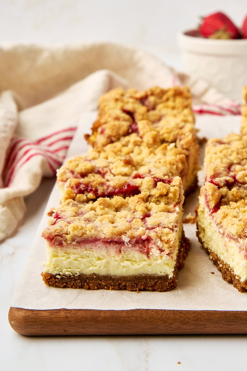a strawberry cheesecake bar on parchment paper with more bars and a bowl of strawberries surrounding