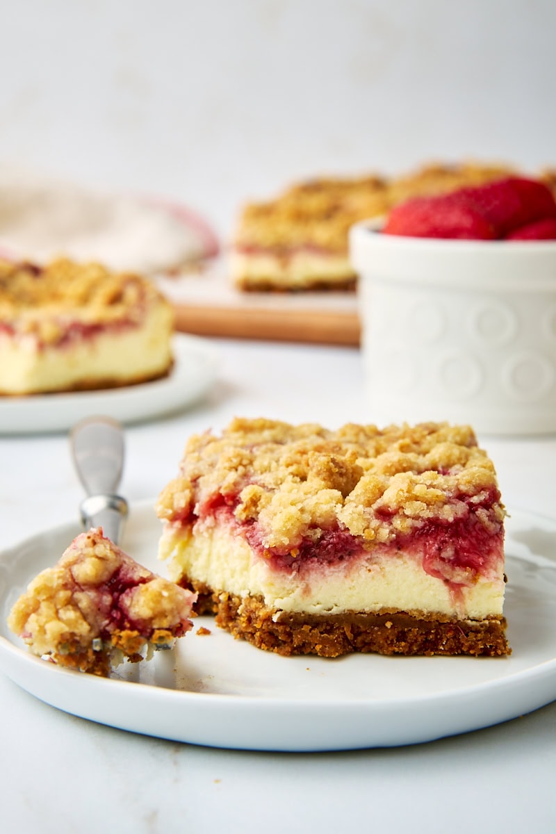 a strawberry cheesecake bar on a white plate with a bite on a fork; more bars and a bowl of fresh strawberries in the background