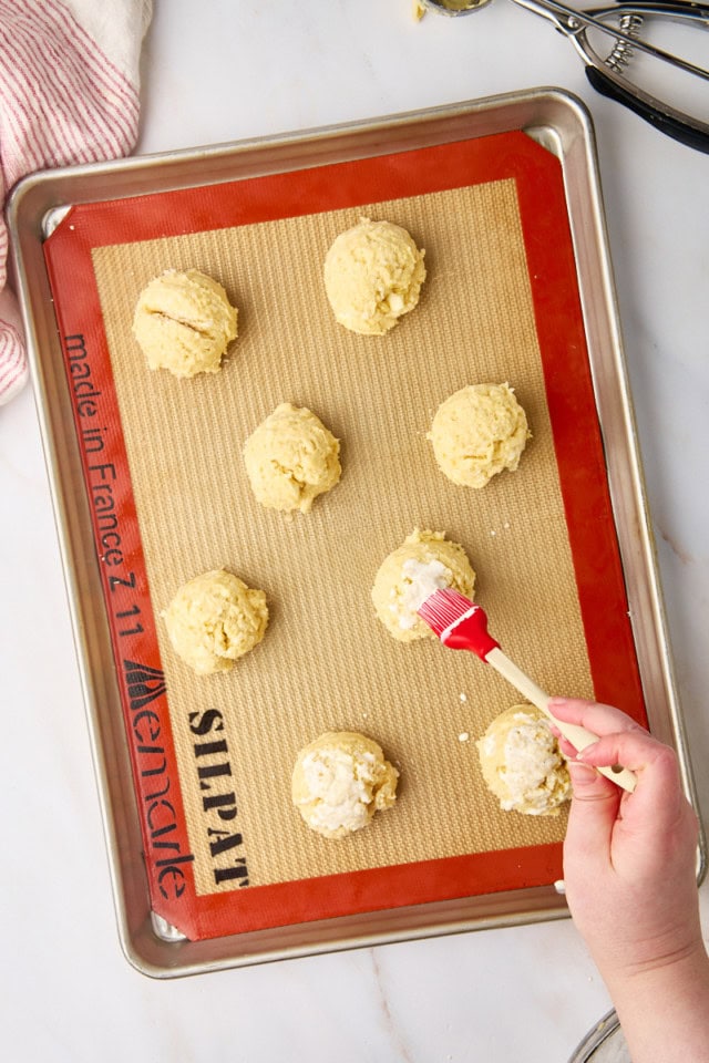 overhead view of biscuit dough on a lined baking sheet with cream being brushed onto them