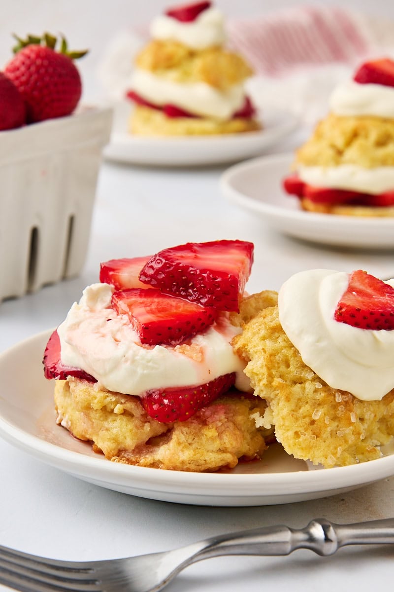 a strawberry shortcake on a white plate with the top set to the side of the plate; more shortcakes and a white basket of strawberries partially visible in the background