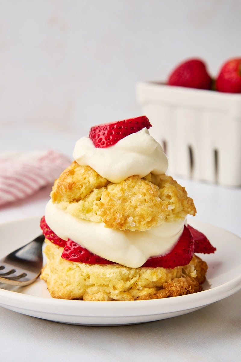 a strawberry shortcake on a white plate with a white basket of strawberries in the background
