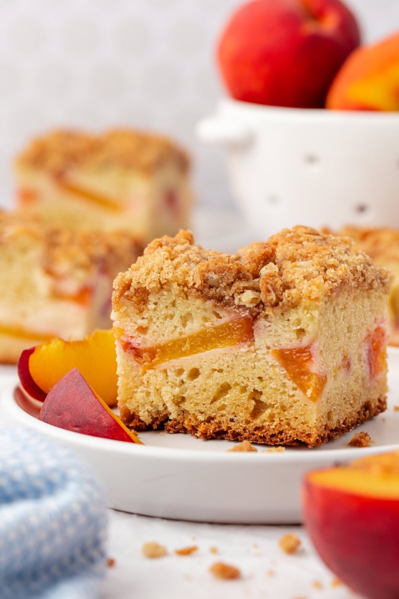 a slice of peach coffee cake on a white plate with more cake and fresh peaches in the background
