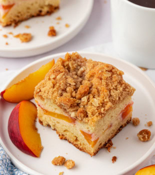a slice of peach coffee cake on a white plate with another slice of cake and a mug of coffee partially visible in the background