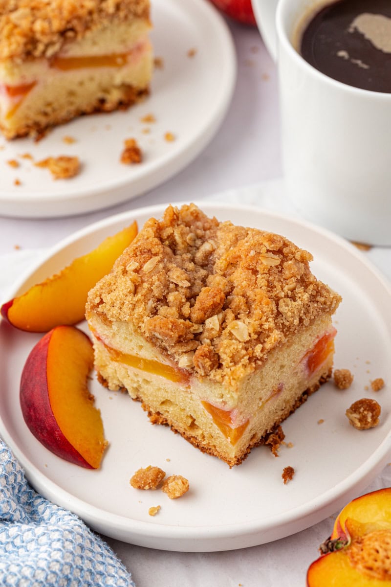 a slice of peach coffee cake on a white plate with another slice of cake and a mug of coffee partially visible in the background