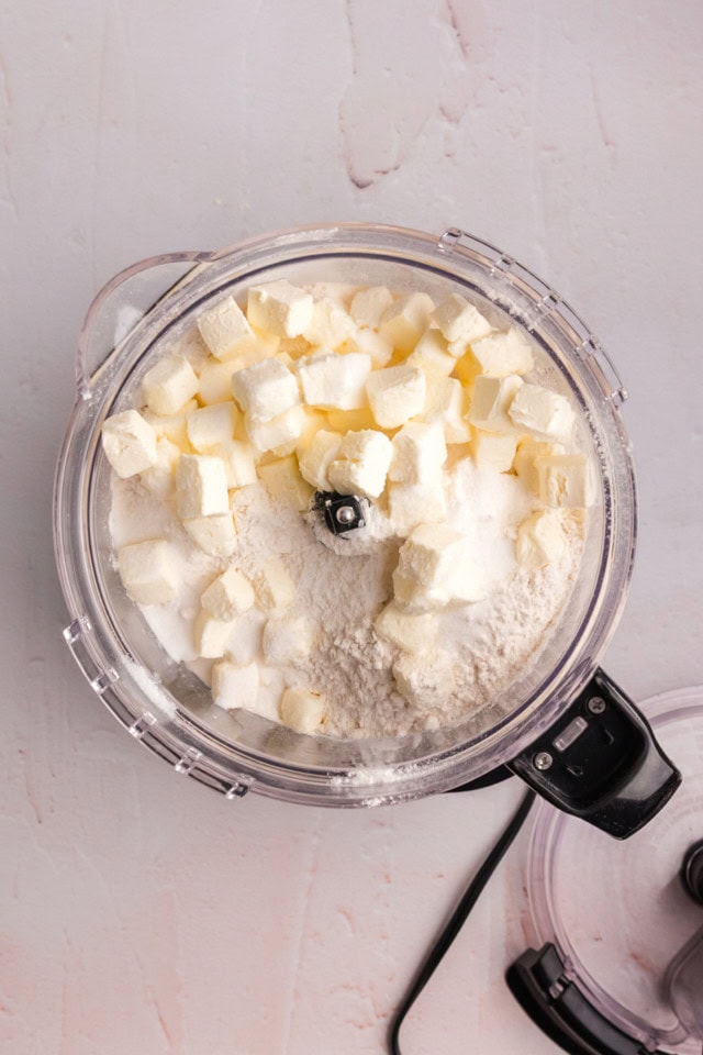 overhead view of butter, cream cheese, flour, sugar, and salt in the bowl of a food processor