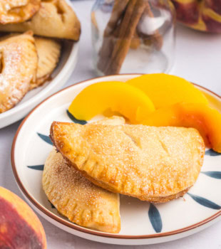 two peach hand pies on a plate with more pies and sliced peaches in the background