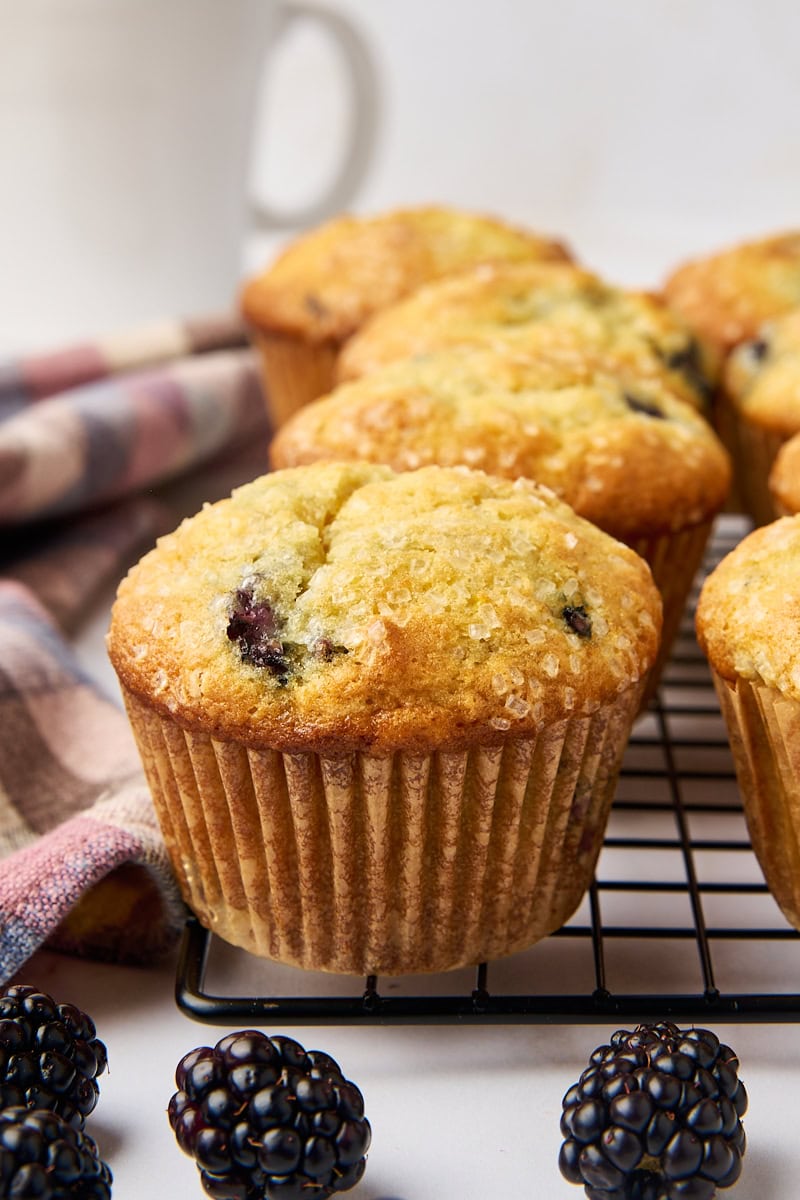 a blackberry muffin on a wire rack along with more muffins, a white coffee mug and fresh blackberries surrounding