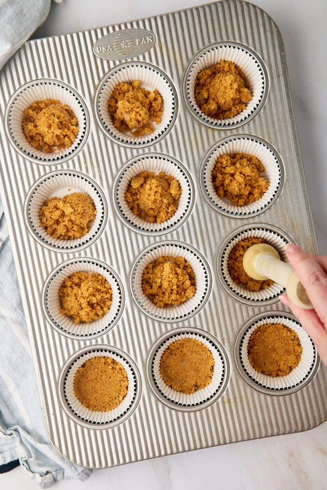 overhead view of graham cracker crust mixture being pressed into the bottoms of lined muffin cups