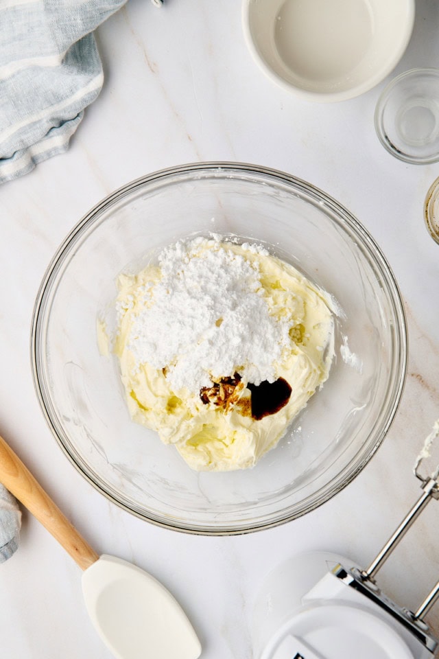 overhead view of confectioners' sugar, vanilla bean paste, and lemon juice added to beaten cream cheese in a glass mixing bowl