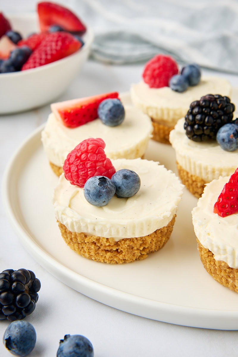 several mini no-bake cheesecakes topped with fresh berries and served on a white plate with more berries in a bowl partially visible in the background