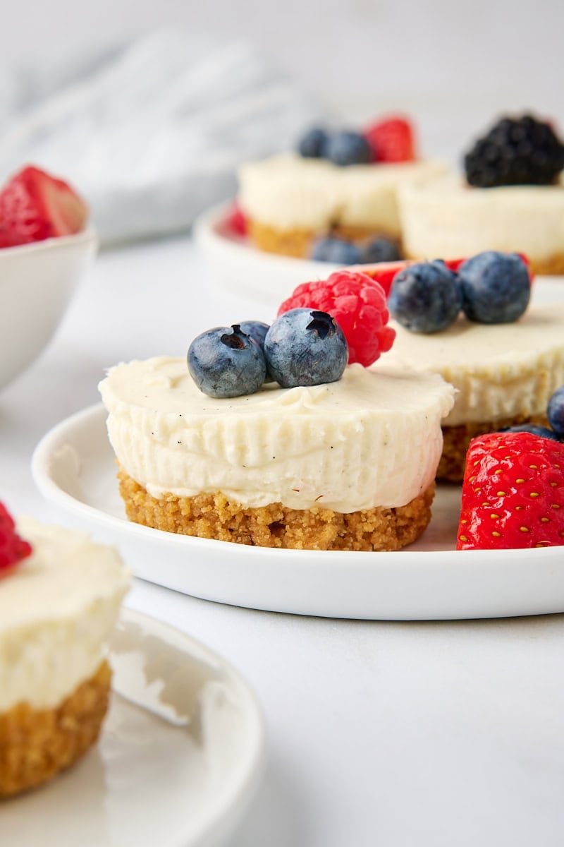 two mini no-bake cheesecakes on a white plate surrounded by more cheesecakes and fresh berries