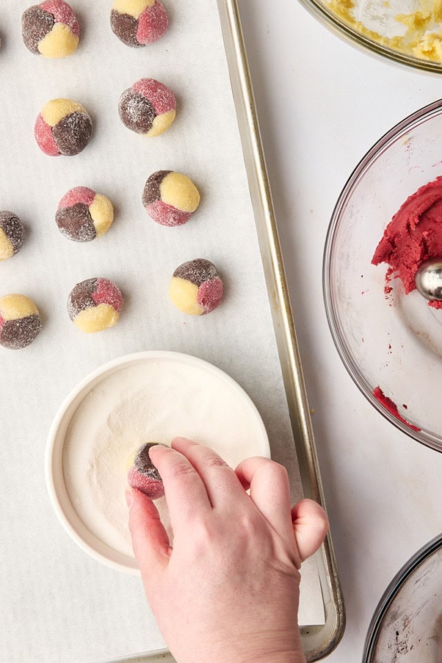 a Neapolitan cookie dough ball being rolled in sugar alongside a pan of more dough balls