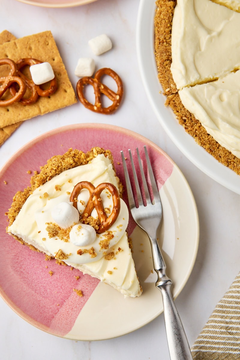 overhead view of a slice of no-bake marshmallow cheesecake topped with whipped cream, mini marshmallows, and pretzels on a pink and white plate with remaining cheesecake partially visible