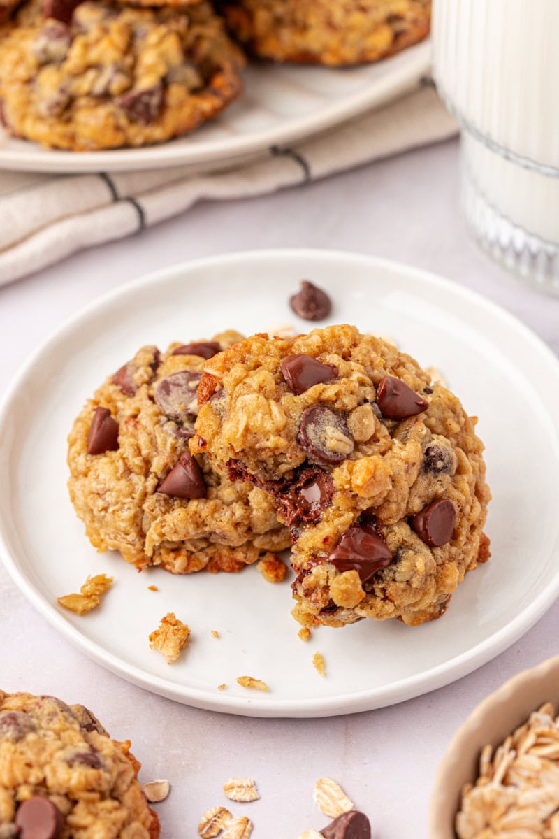 two brown sugar oatmeal chocolate chip cookies on a white plate with more cookies surrounding