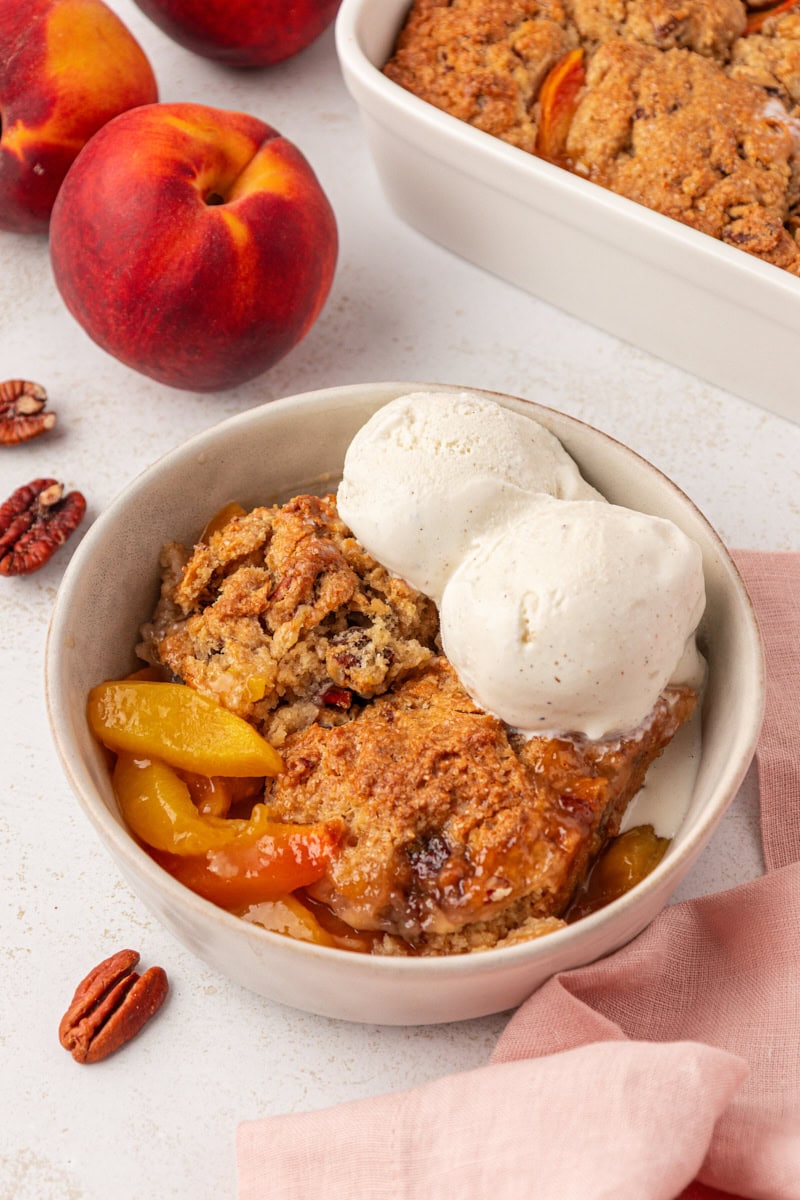 ginger peach cobbler topped with two scoops of ice cream in a bowl with the remaining cobbler and fresh peaches in the background