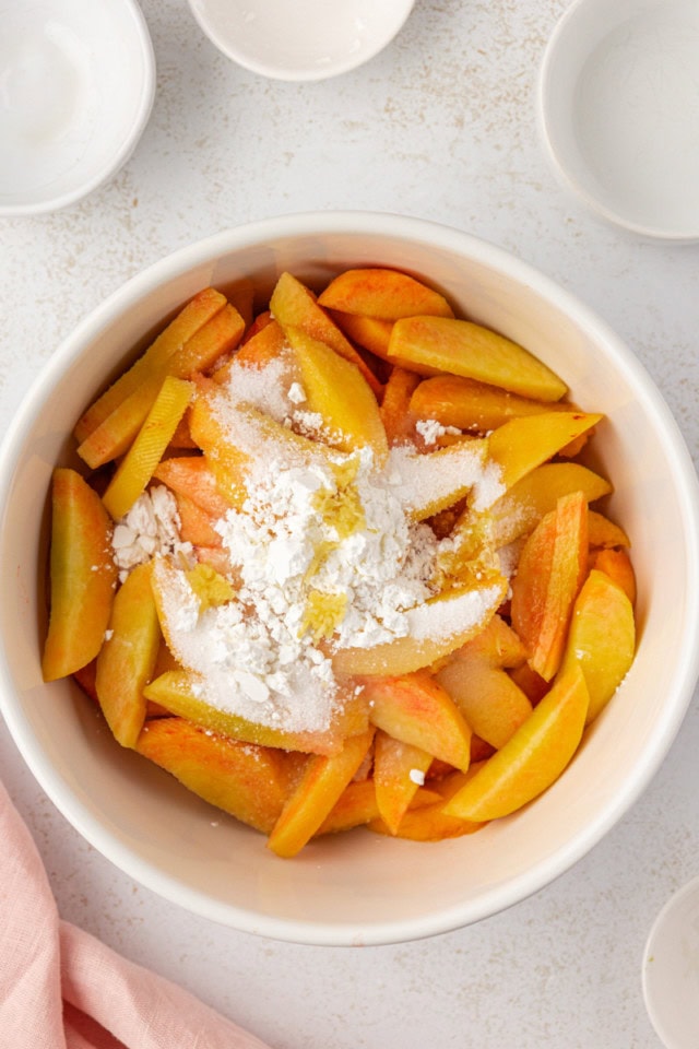 overhead view of peaches, sugar, lemon juice, ginger, and cornstarch in a white mixing bowl