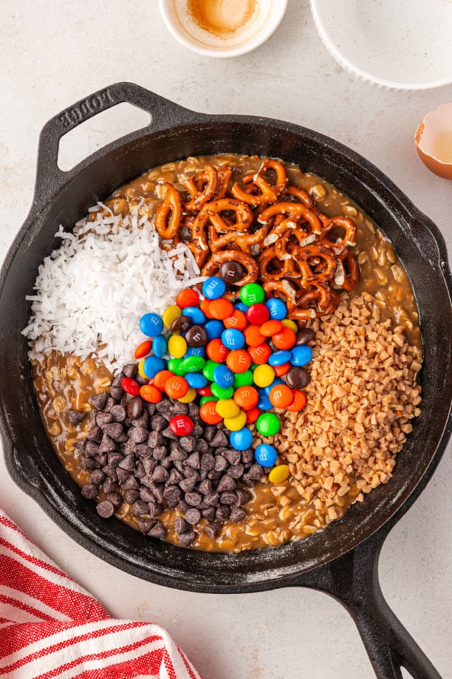 overhead view of chocolate chips, M&Ms, pretzels, and more add-ins added to cookie dough in a cast iron skillet