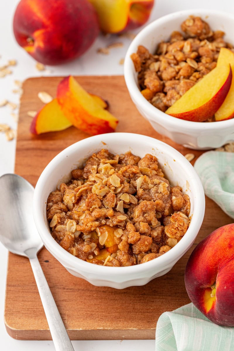 a serving of peach crisp on a wooden board with another serving and fresh peaches in the background
