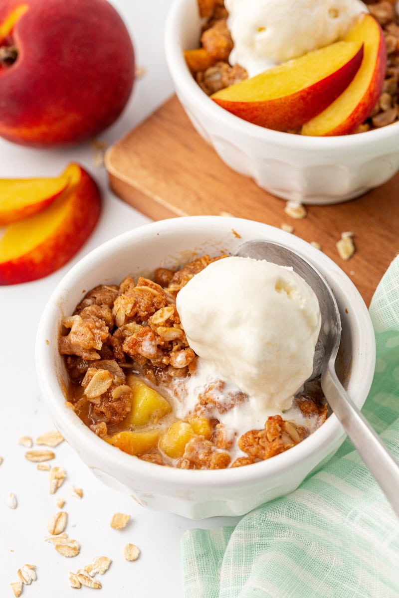 peach crisp topped with ice cream in a white bowl with another serving and fresh peaches in the background